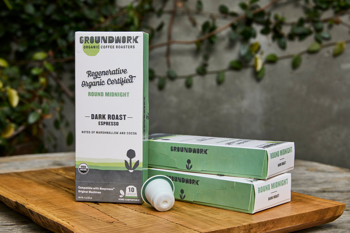 Groundwork Coffee Regenerative Organic Certified® Round Midnight - Nespresso® pods on a wooden board, with plants and a gray wall in the background.