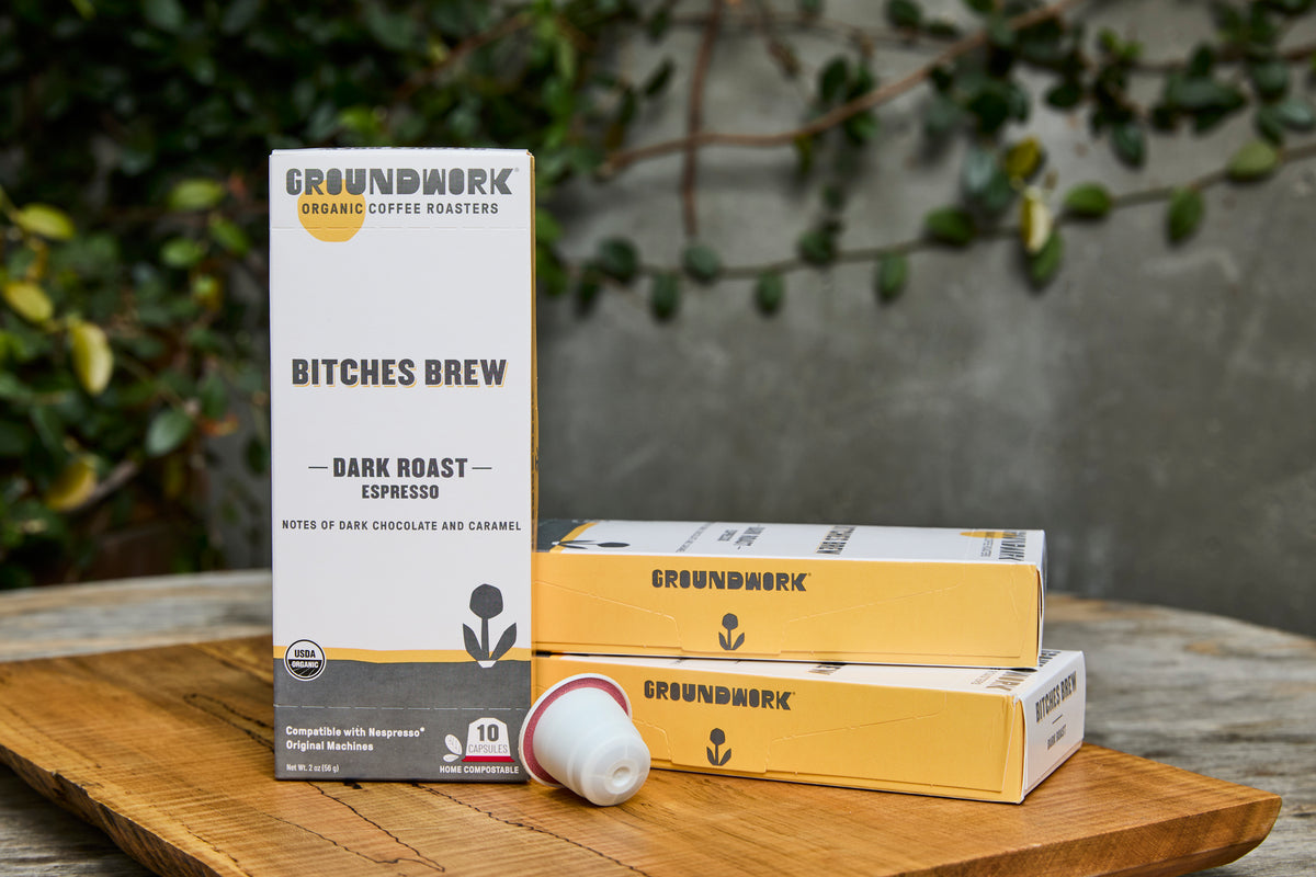 A box and two packets of Groundwork Coffee Bitches Brew - Nespresso® pods on a wooden surface, with greenery in the background.