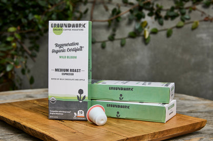 Groundwork Coffee’s Regenerative Organic Certified® Wild Bloom - Nespresso® pods, two boxes and a pod, on a wooden board outdoors among leafy plants.
