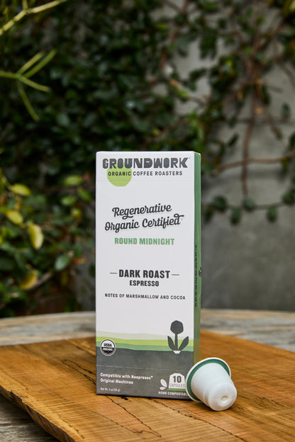 A box of Groundwork Coffee Regenerative Organic Certified® Round Midnight - Nespresso® pods sits outdoors on wood with greenery behind it; one pod is placed next to the box.