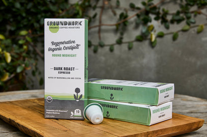 Groundwork Coffee Regenerative Organic Certified® Round Midnight - Nespresso® pods on a wooden board, with plants and a gray wall in the background.