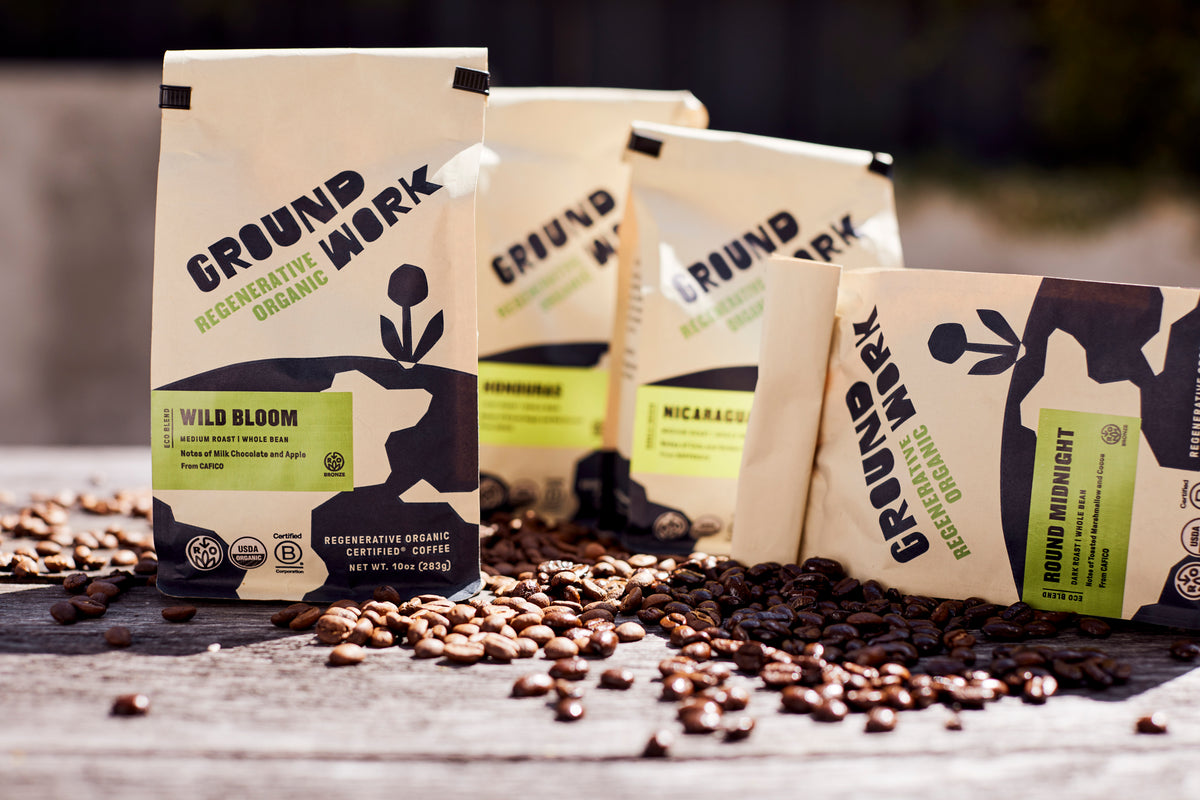 Groundwork Regenerative Organic Certified® coffee bags in cream-colored packaging with black and green design, arranged outdoors on a wooden surface with scattered roasted coffee beans. Visible blends include Wild Bloom and Round Midnight, along with single-origin coffees from Honduras and Nicaragua.