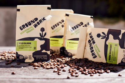Groundwork Regenerative Organic Certified® coffee bags in cream-colored packaging with black and green design, arranged outdoors on a wooden surface with scattered roasted coffee beans. Visible blends include Wild Bloom and Round Midnight, along with single-origin coffees from Honduras and Nicaragua.