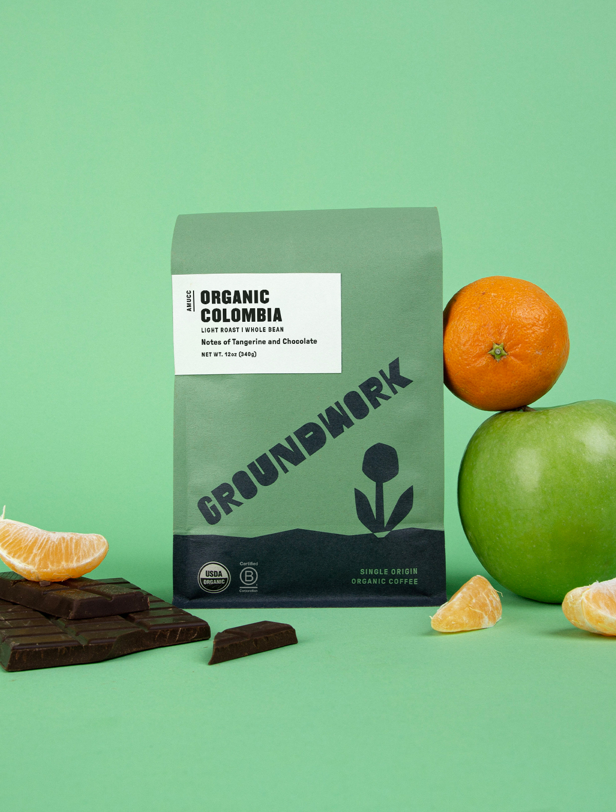A green bag of Groundwork Coffees Colombia: AMUCC Fair Trade is shown with an orange, green apple, milk chocolate, and orange slices on a green background.