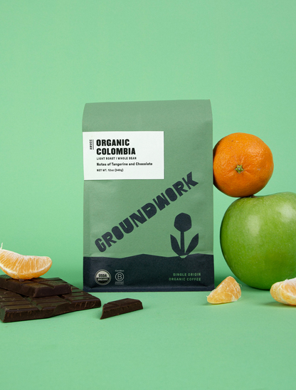 A green bag of Groundwork Coffees Colombia: AMUCC Fair Trade is shown with an orange, green apple, milk chocolate, and orange slices on a green background.