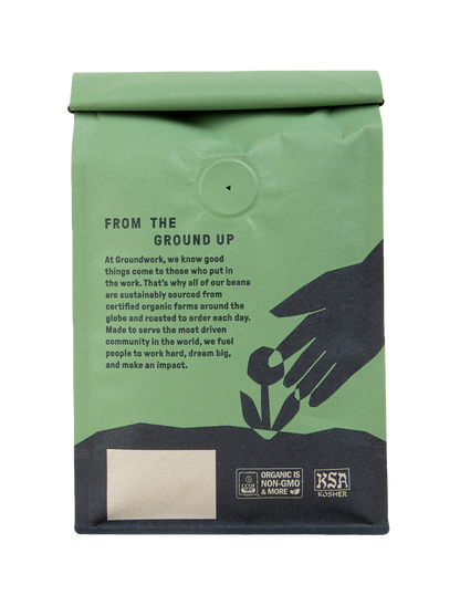 A green bag of Groundwork Coffee’s Colombia: AMUCC Fair Trade features black hands cradling a sprout. Light roast with tangerine, milk chocolate notes. Highlights organic, sustainable, Kosher, non-GMO.