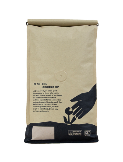 A brown bag of Groundwork Coffee’s Colombia: AMUCC Fair Trade features black graphics of a hand planting a seedling, with Light Roast and fair trade certification logos displayed.
