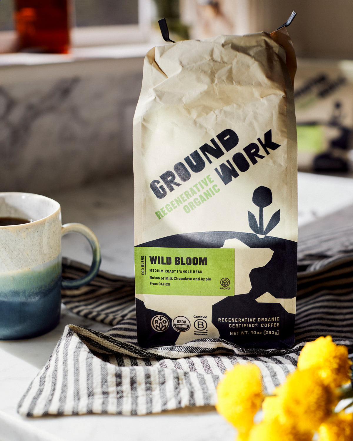 A bag of Groundwork Coffee Regenerative Organic Certified® Wild Bloom sits on a striped cloth beside a mug and yellow flowers, with sunlight streaming in from the left.