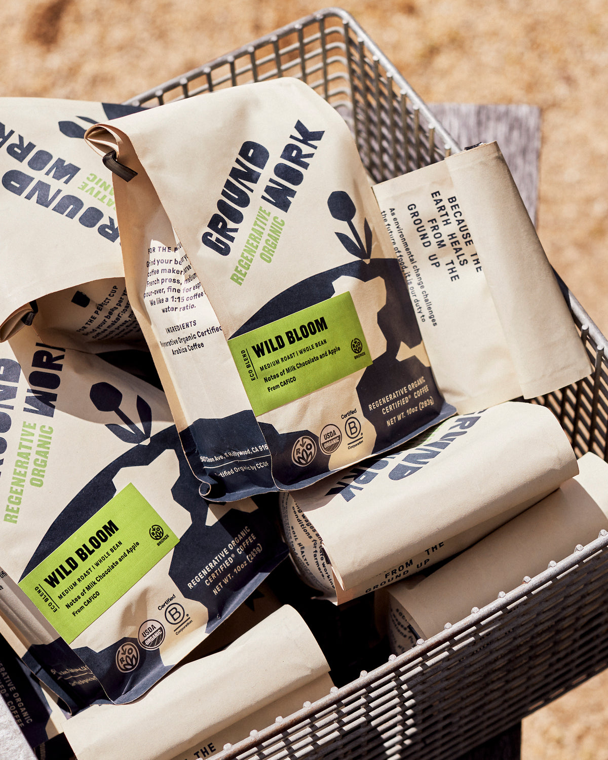 Several bags of Groundwork Coffee Regenerative Organic Certified® Wild Bloom, a medium roast with milk chocolate and apple notes, are stacked in a wire basket outdoors.