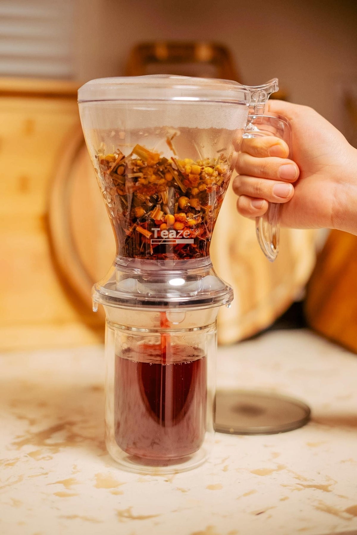 A hand uses the Groundwork Coffee Teaze Tea Infuser filled with loose leaf tea, letting brewed tea drain into a glass mug below.