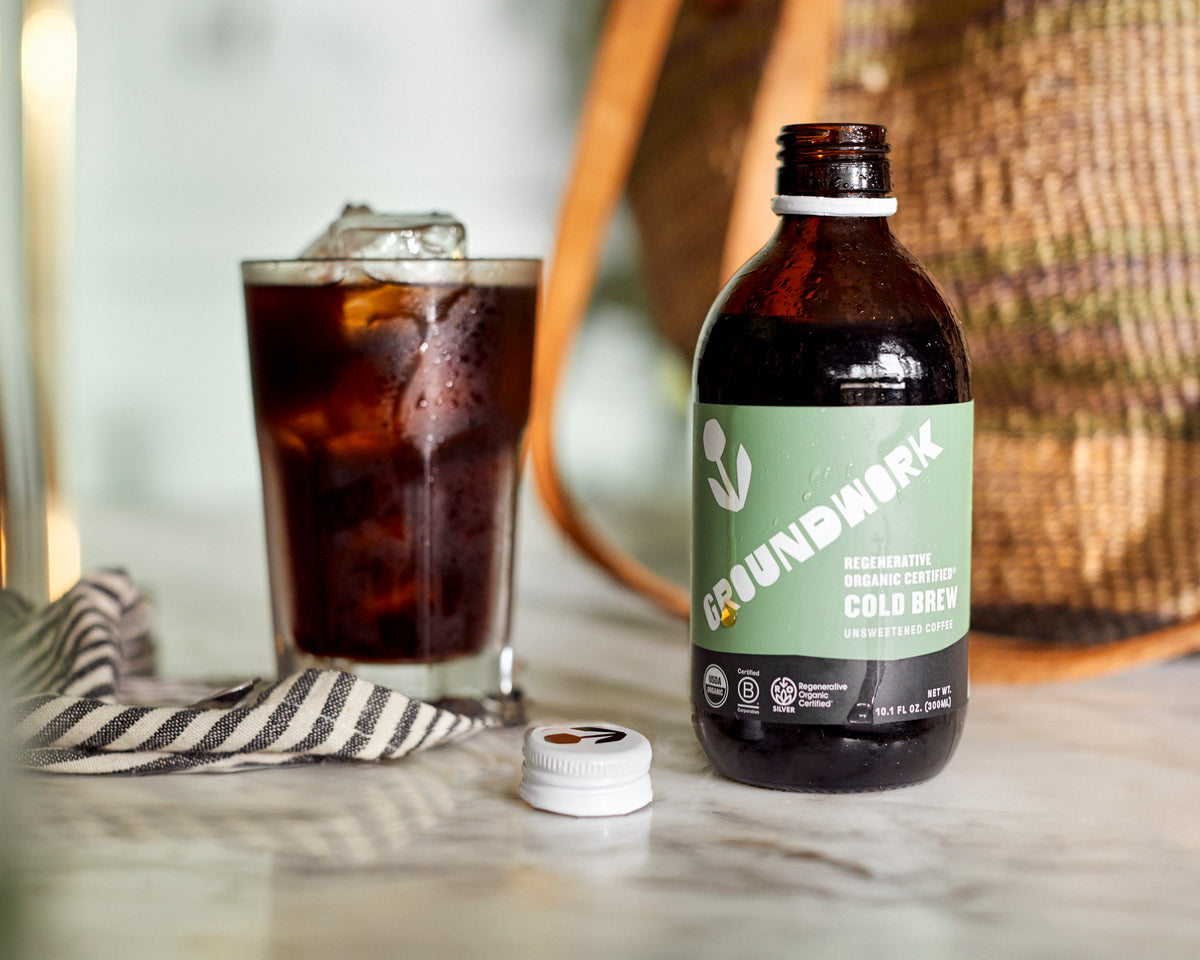 A bottle of Groundwork Coffee Co Regenerative Organic Certified® Cold Brew sits open beside a glass of iced coffee and a striped napkin on marble.