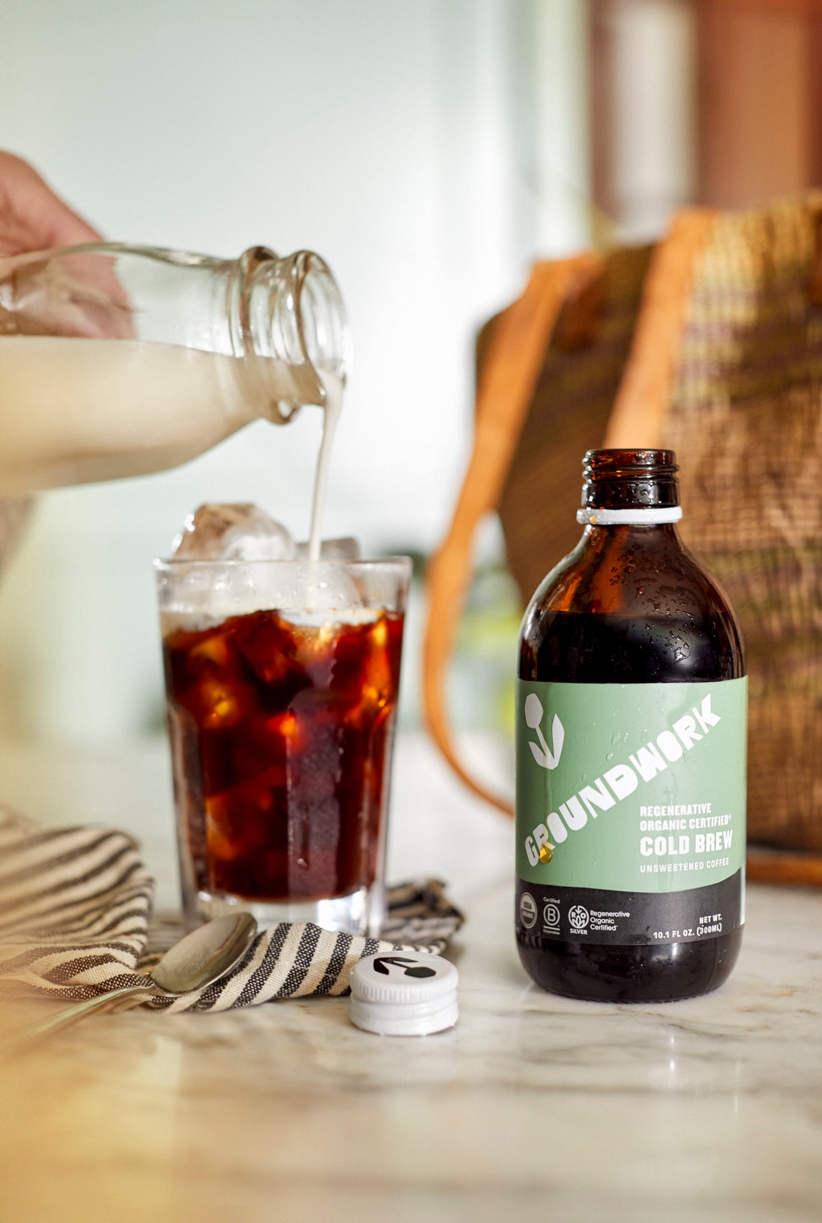 A hand pours milk into iced coffee next to a Regenerative Organic Certified® Cold Brew by Groundwork Coffee Co on a marble countertop.