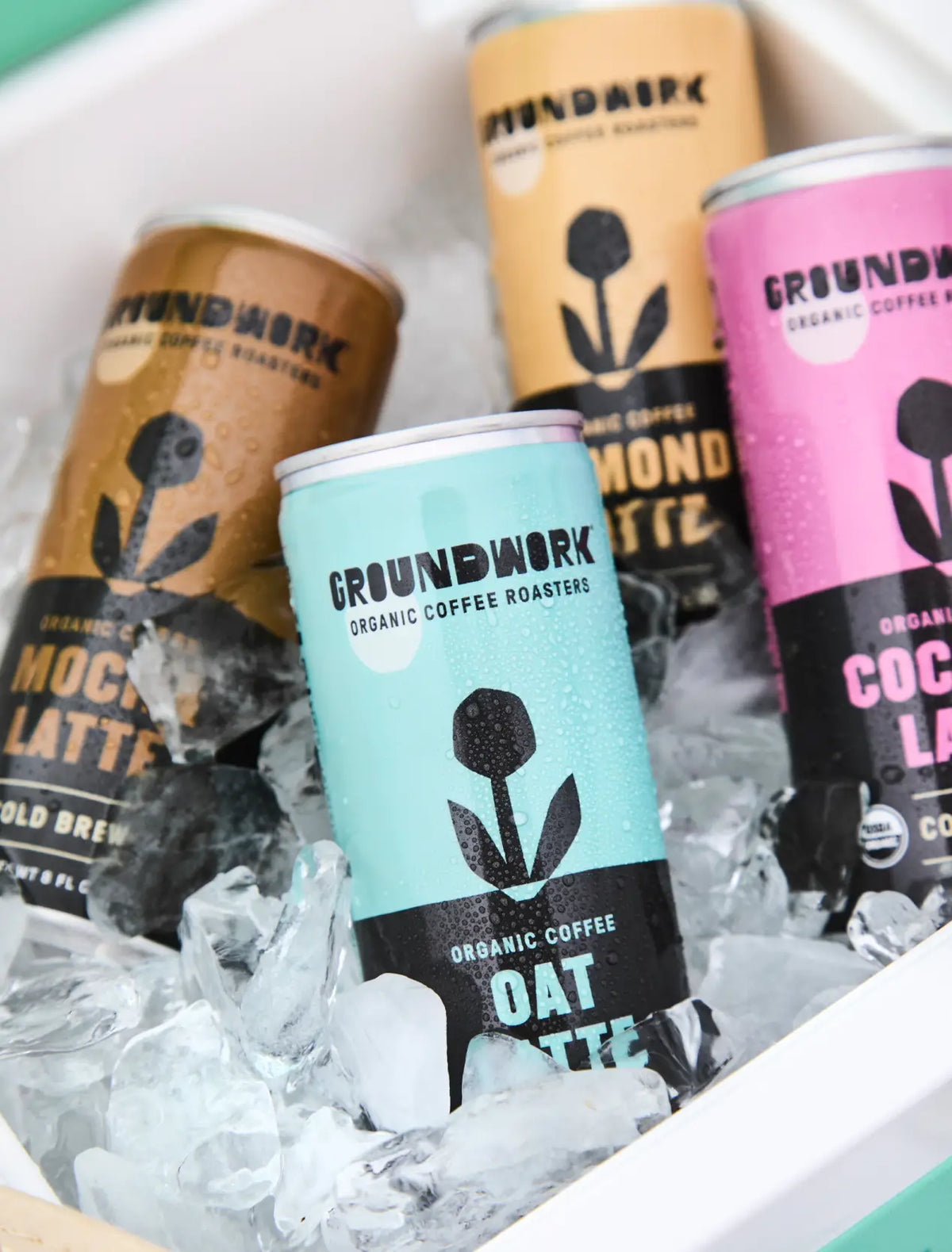 Four cans of Groundwork Coffee Organic Cold Brew Latte in assorted flavors sit in ice, condensation beading on each can.