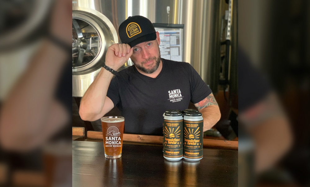 A man in a black Santa Monica Brew Works shirt and hat sits at a bar with a pint of beer and two cans, posing with one hand touching his hat. Brewery equipment is in the background.