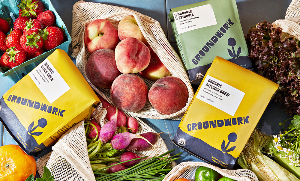 Assorted fresh fruits and vegetables in mesh bags, with three yellow and green Groundwork coffee bags placed among them.