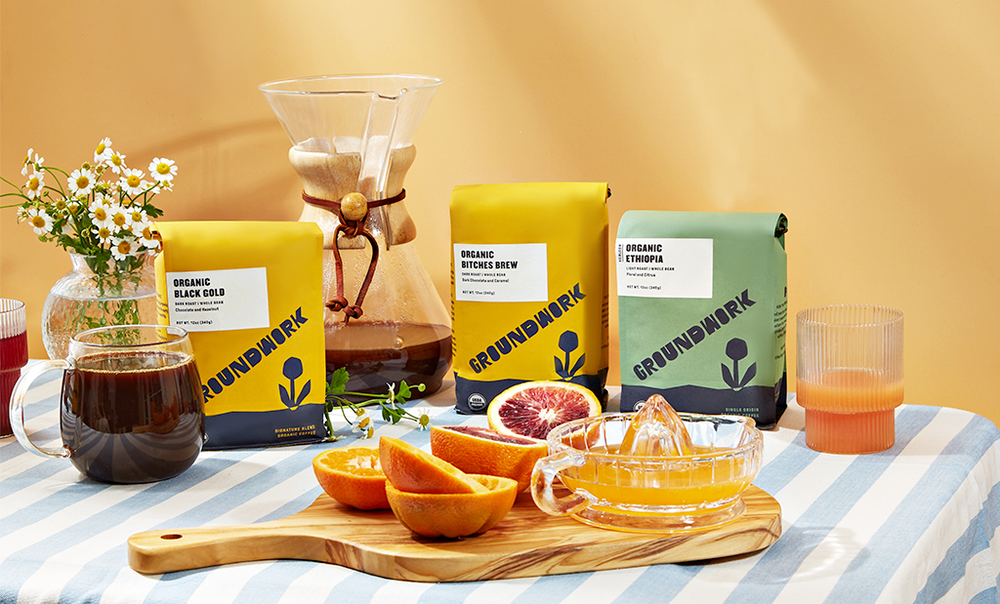 Coffee bags, a Chemex pot, mugs, sliced oranges, a juicer, and flowers arranged on a striped tablecloth, with a warm, orange background.