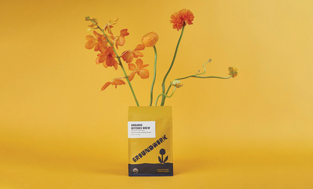 A yellow bag of Groundwork Organic Diverse Brew coffee with orange flowers arranged to look like they are growing from the bag, set against a matching yellow background.