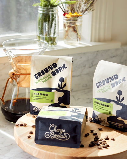 A bag of Groundwork Coffee’s Regenerative Organic Certified® Round Midnight sits on a wooden board with cocoa-hued beans and a Chemex by a sunny window.