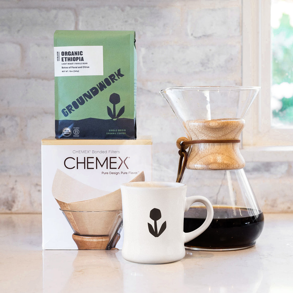 The Chemex Starter Kit from Groundwork Coffee, featuring a brewer, filters, coffee, and a white mug, sits on a counter by the window.