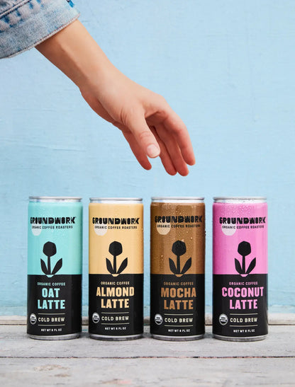 A hand reaches for four cans of Groundwork Coffee Organic Cold Brew Latte—Oat, Almond, Mocha, and Coconut—against a blue wall.