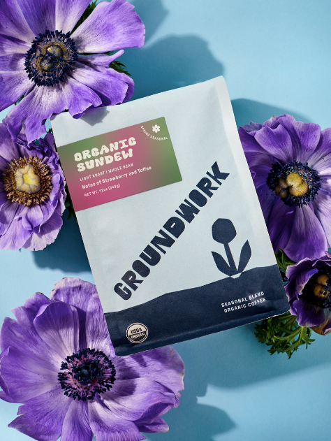 A bag of Groundwork Coffee's Seasonal Select: Sundew rests on a blue surface, surrounded by purple flowers. Its label features delicate strawberry and toffee notes.