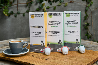 A cup of espresso sits on a wooden tray next to three boxes of Groundwork Coffee Nespresso® Variety Pack pods, with greenery in the background.