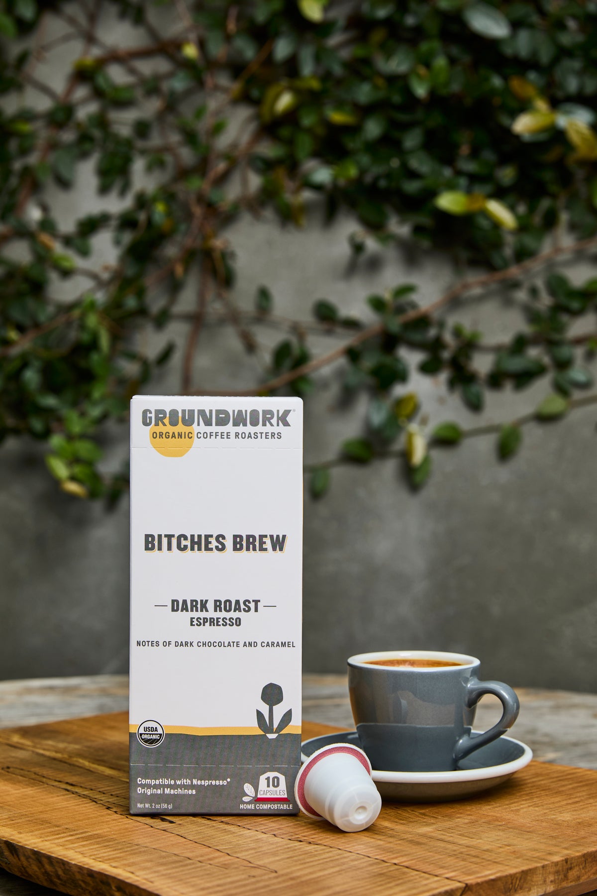 A box of Groundwork Coffee Bitches Brew - Nespresso® pods sits on a wooden table beside a grey cup, with leafy plants in the blurred background.