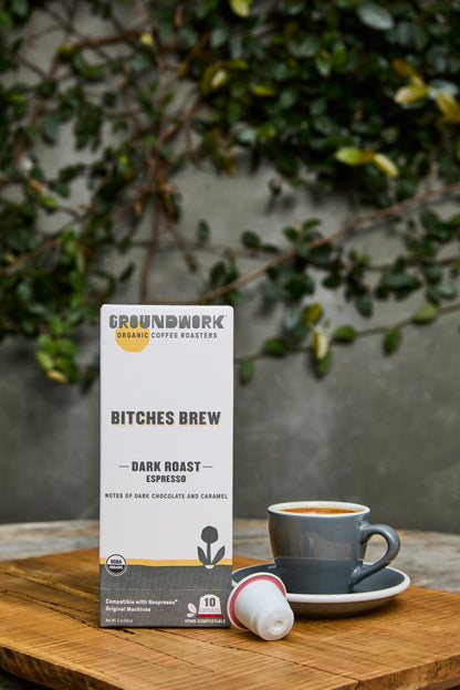 A box of Groundwork Coffee Bitches Brew - Nespresso® pods sits on a wooden table beside a grey cup, with leafy plants in the blurred background.