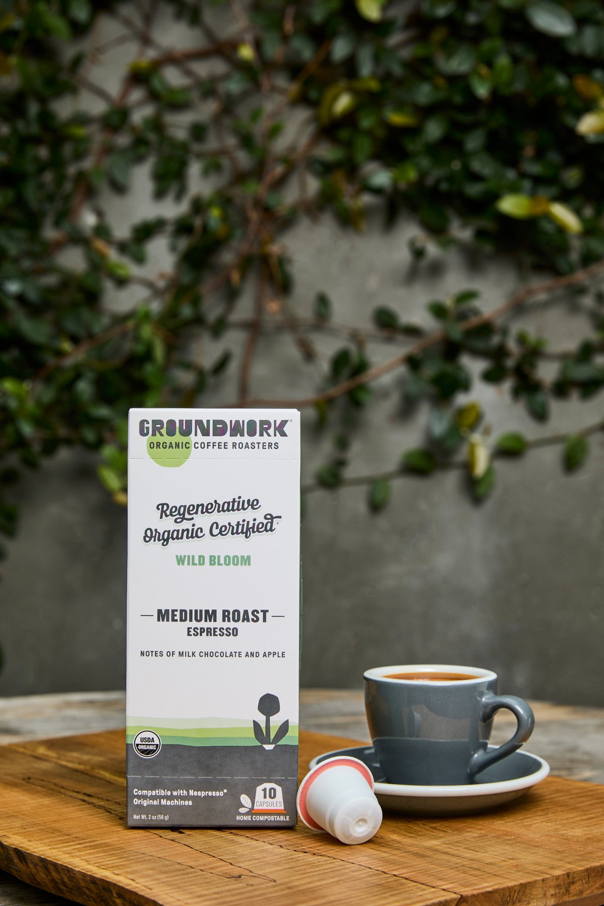 A box of Groundwork Coffee's Regenerative Organic Certified® Wild Bloom - Nespresso® sits by a coffee cup and pod on a wooden table with green leaves in the background.