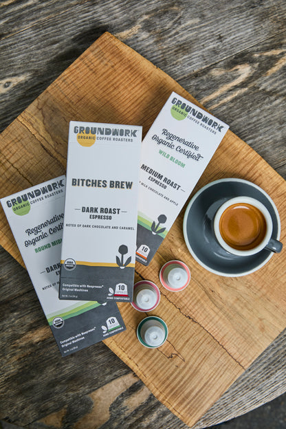 A cup of espresso sits on a wooden board beside three Groundwork Coffee Nespresso® Variety Pack boxes with certified compostable pods.