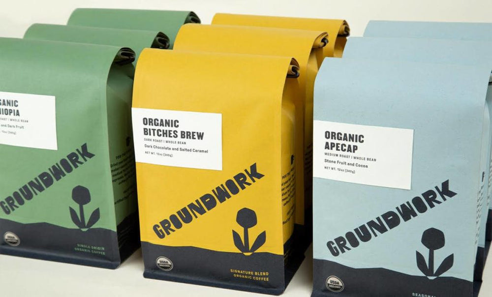 Three flavors of Groundwork organic coffee bags—Ethiopia (green), Bitches Brew (yellow), and Ape Cap (blue)—are arranged side by side on a white surface.