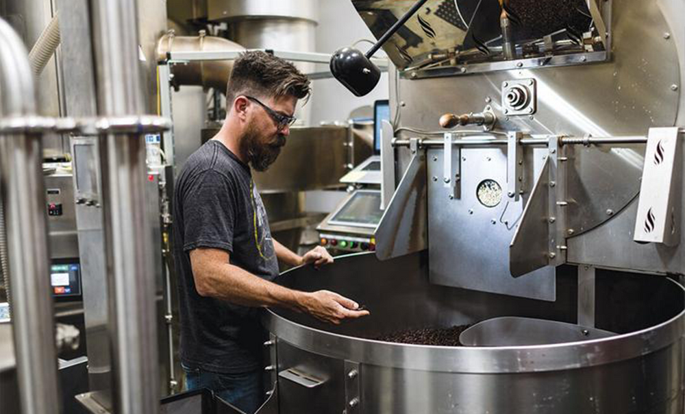 A man with a beard inspects roasted coffee beans in a large industrial roasting machine at a coffee production facility.