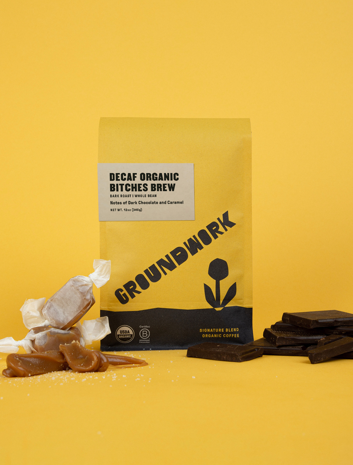 A yellow bag of Groundwork Coffee’s Decaf Bitches Brew stands on a yellow background, surrounded by caramels and dark chocolate pieces.