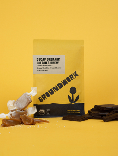 A yellow bag of Groundwork Coffee’s Decaf Bitches Brew stands on a yellow background, surrounded by caramels and dark chocolate pieces.