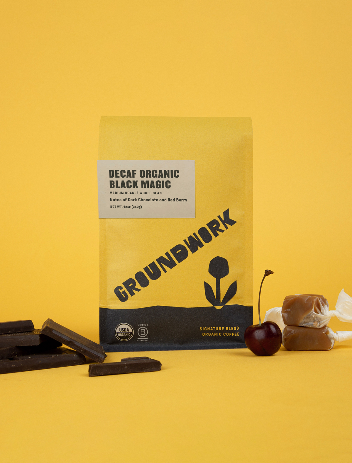A 12oz yellow bag of Groundwork Decaf Black Magic Espresso stands upright against a yellow background, surrounded by dark chocolate, caramel, and a red cherry.