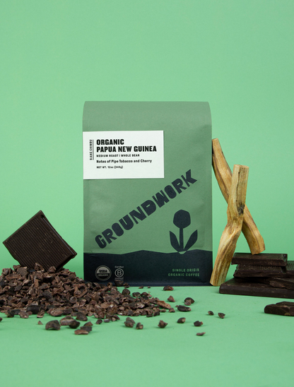 A green bag of Groundwork Coffee Stores Papua New Guinea: Siane Chimbu is surrounded by dark chocolate pieces and cocoa nibs on a green background.