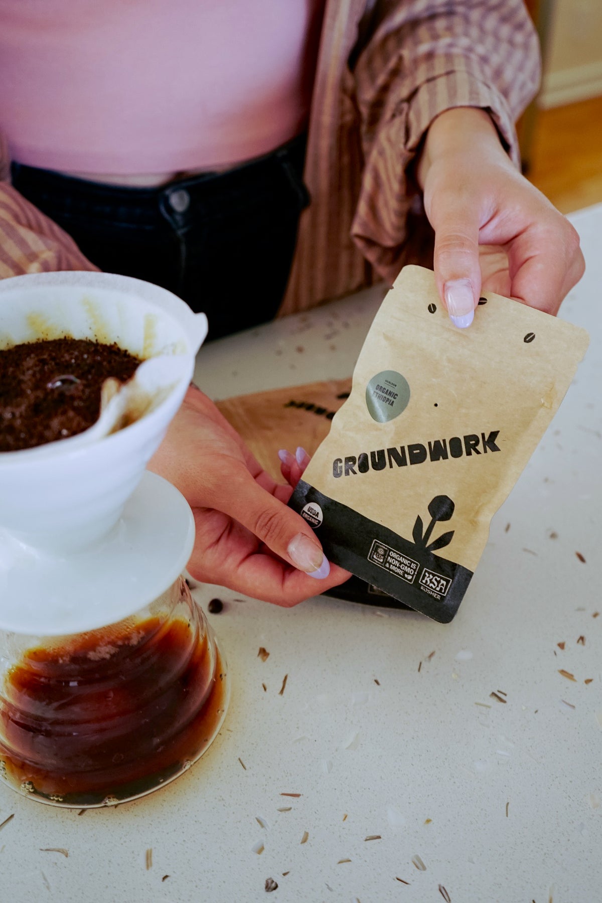 A person holding a small tan and black, 2oz Groundwork Organic Coffee Roasters sample pouch near a white ceramic pour-over coffee dripper and a glass carafe of brewed coffee on a countertop. 