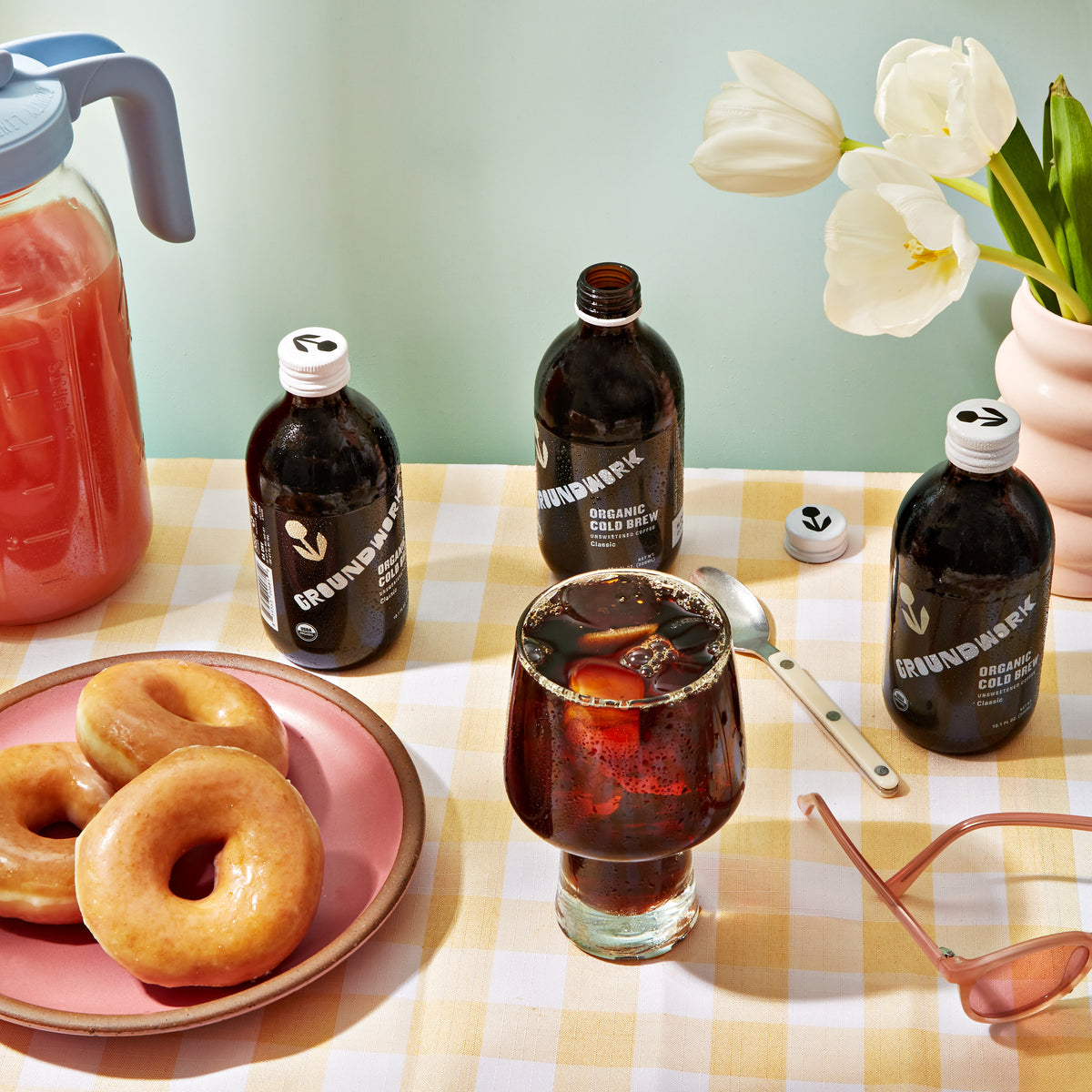 A table with glazed donuts, a glass of Organic Classic Cold Brew by Groundwork Coffee Co, bottles, white tulips in a vase, and pink sunglasses on a yellow checked tablecloth.