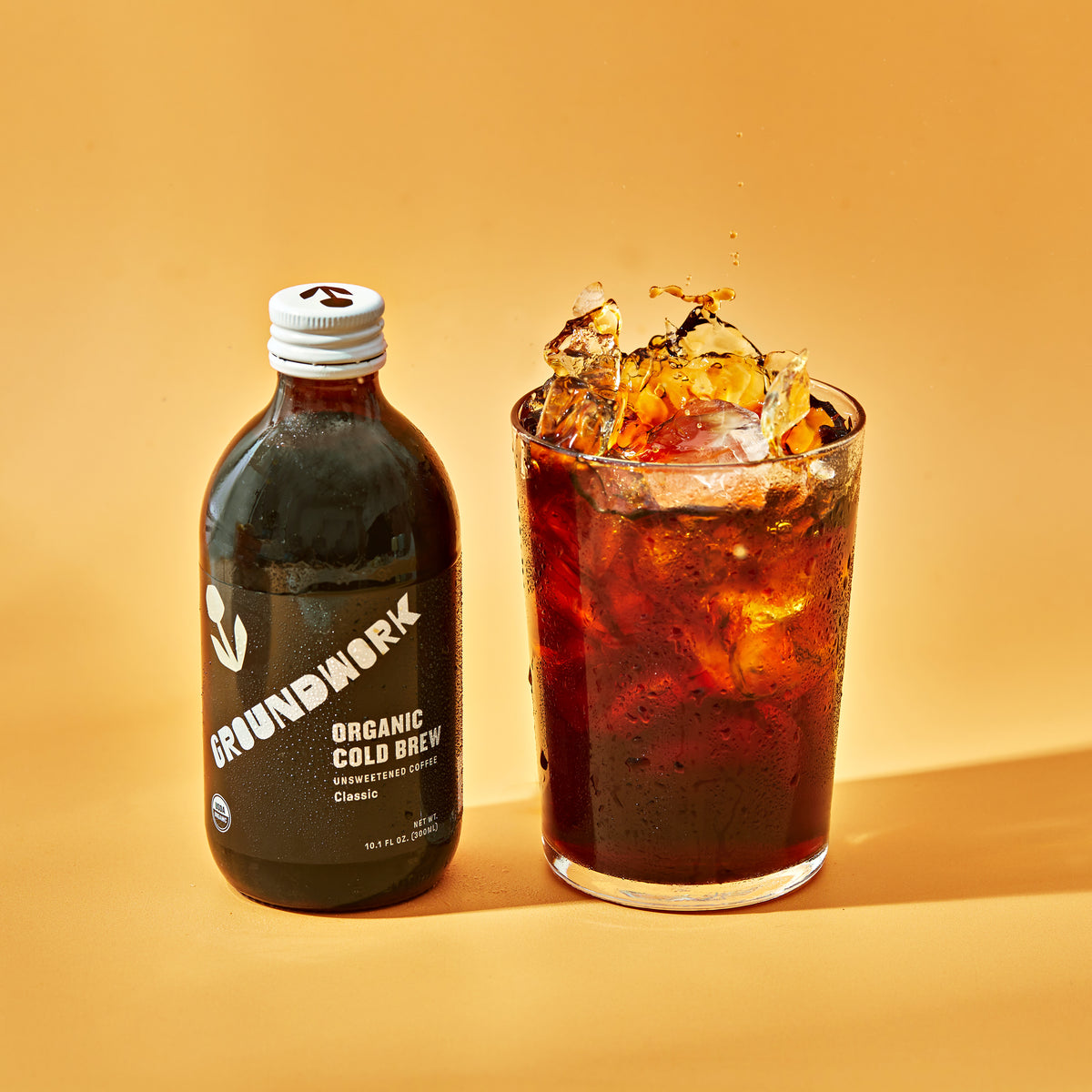 A bottle of Groundwork Coffee Co Organic Classic Cold Brew sits by a glass of iced coffee against a bold orange background.