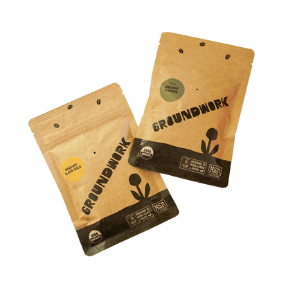 Two small, kraft paper 2oz sample coffee pouches from Groundwork Organic Coffee Roasters displayed on a white background. 
One pouch is labeled “Organic Black Gold” with a small, yellow sticker, and the other is labeled “Organic Ethiopia” with a small, green sticker. 
Both bags feature the Groundwork logo, USDA Organic and KSA Kosher certifications, and a black flower graphic at the bottom.